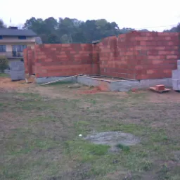 Wznoszenie murów z czerwonej cegły na fundamencie betonowym, widok na budowę domu jednorodzinnego w trakcie realizacji, sterta bloczków obok.